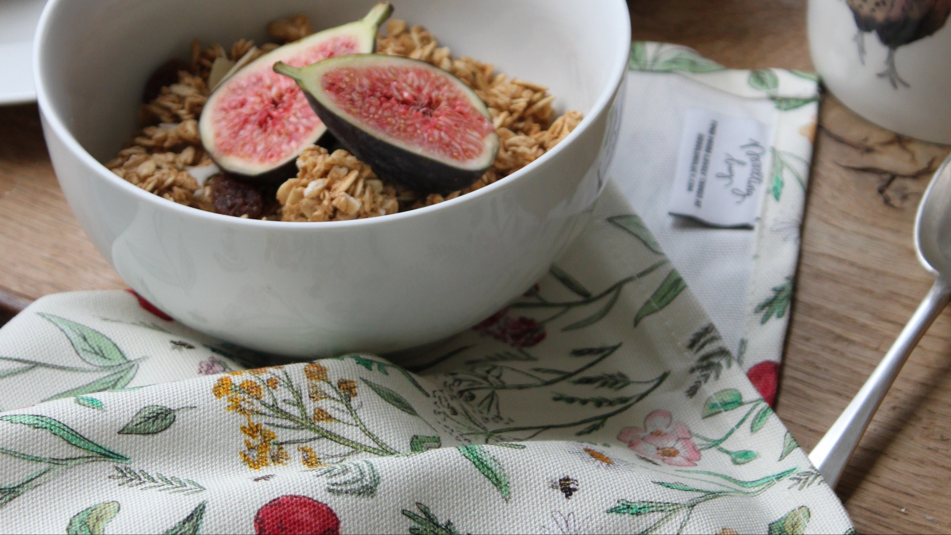 A white ceramic bowl filled with granola and sliced fresh figs, set on a floral napkin on a wooden table. A white mug with a pheasant illustration is visible on the right, along with blueberries in a bowl in the background.